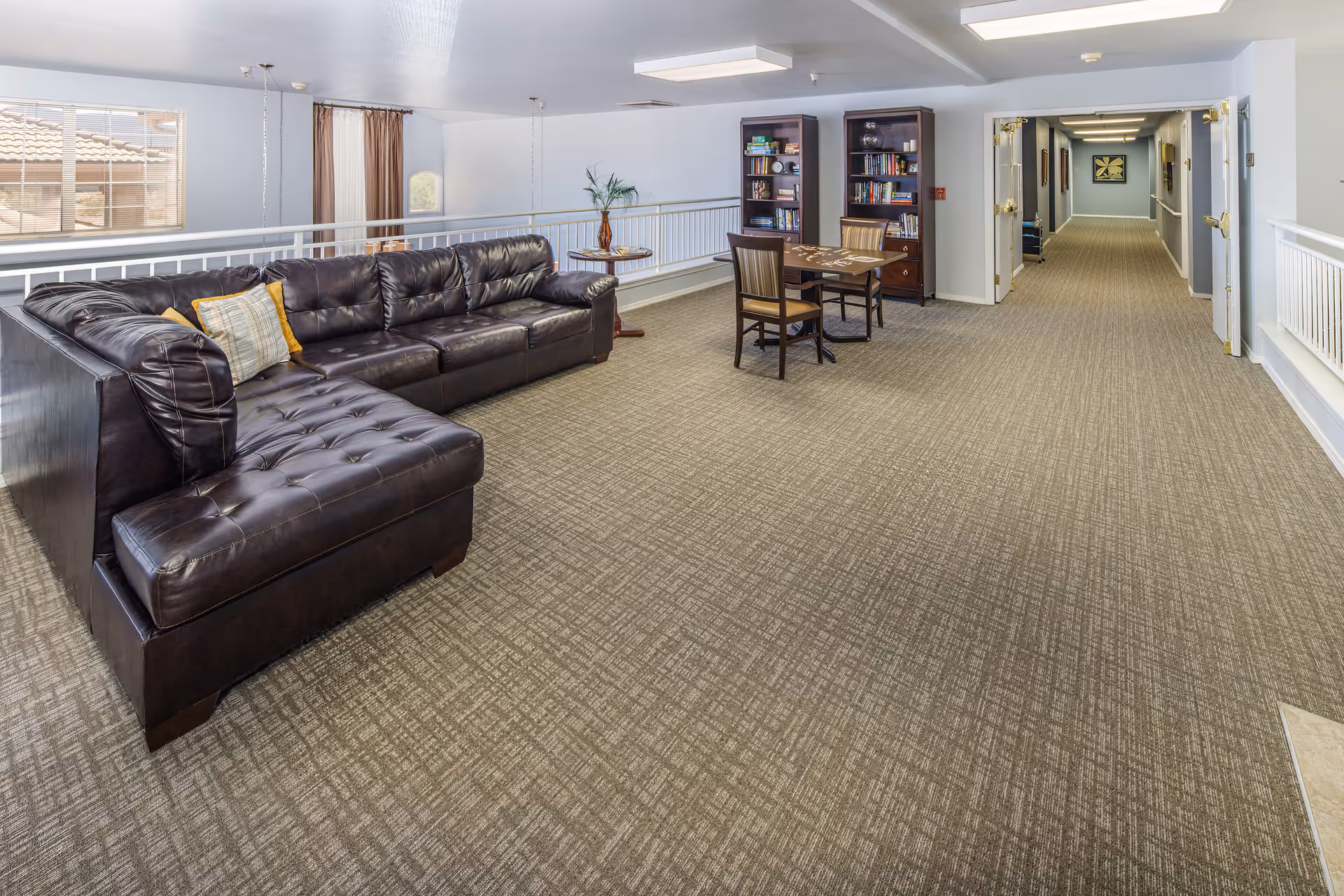 A spacious interior common area with a large dark brown leather sectional sofa adorned with pillows, a small table with two chairs, and two bookshelves filled with books and decorative items. The area has beige patterned carpet, light blue walls, and a hallway leading to other rooms. Large windows allow natural light to enter the space.