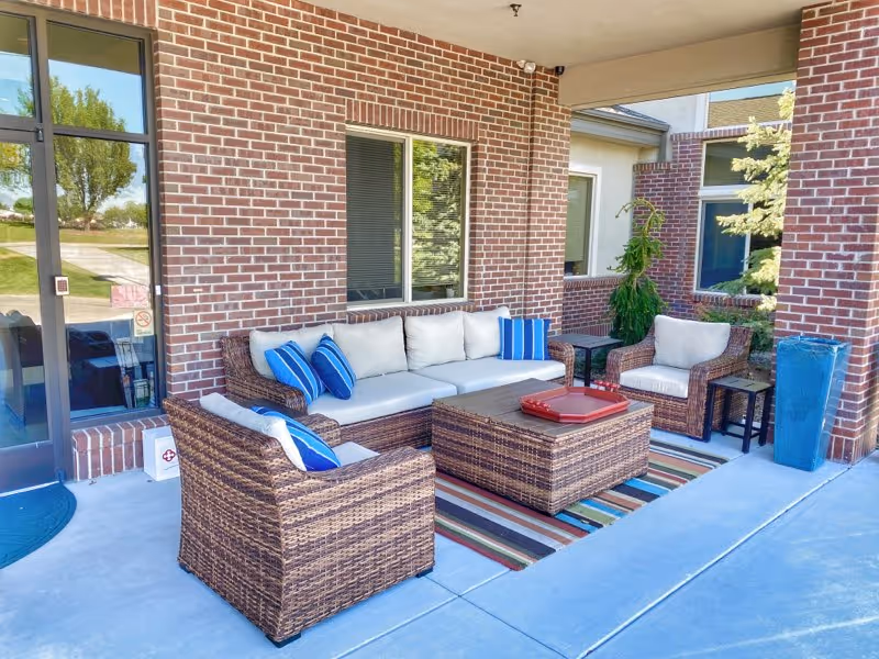 Outdoor seating area with wicker furniture including a sofa, two armchairs, and a coffee table on a striped rug. The seating has white cushions and blue accent pillows. The area is next to a brick wall with windows and a glass door, with some greenery visible nearby.