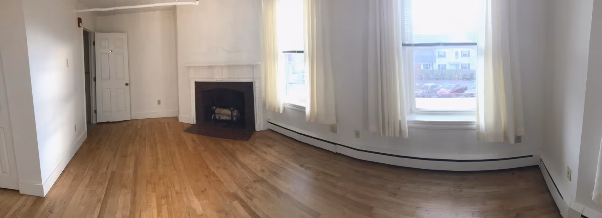 Empty room with light wooden flooring, white walls, a white fireplace, two large windows with white curtains, and a white door slightly ajar.