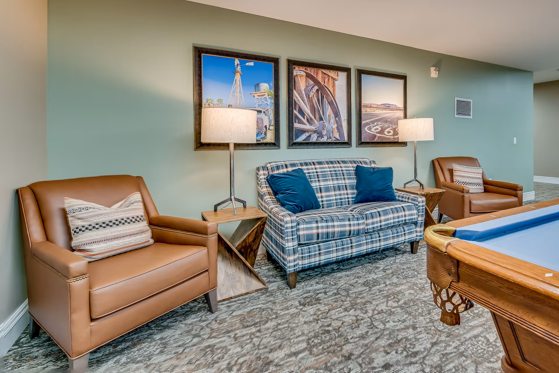 A cozy sitting area in a senior living facility featuring a plaid upholstered loveseat with two blue pillows, flanked by two brown leather armchairs with patterned cushions. Between the loveseat and each armchair are modern wooden side tables with lamps. Above the loveseat are three framed pictures depicting rustic scenes including a windmill, a wagon wheel, and a Route 66 road sign. Part of a pool table is visible in the foreground on the right side.