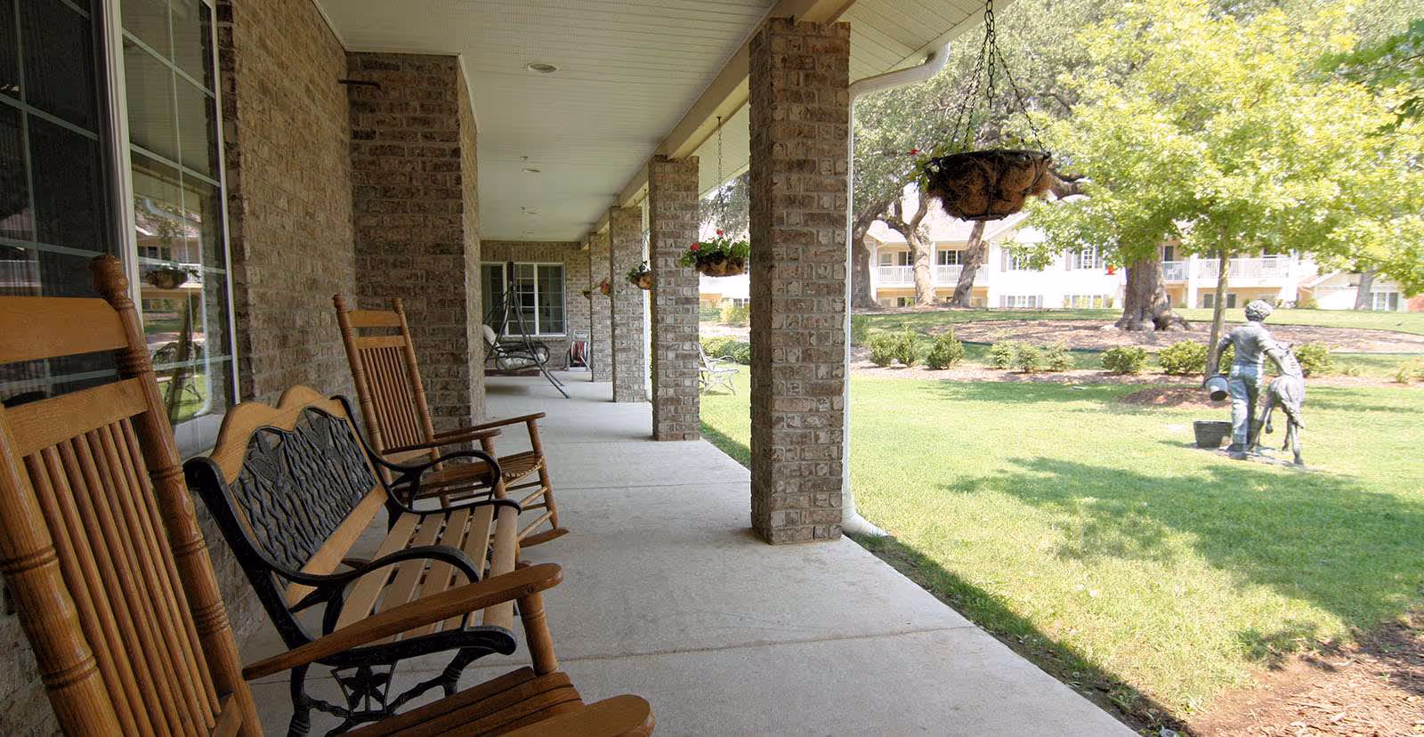 Covered outdoor patio area with wooden rocking chairs and a bench along a brick wall, overlooking a green lawn with trees and a statue of a person with a dog.