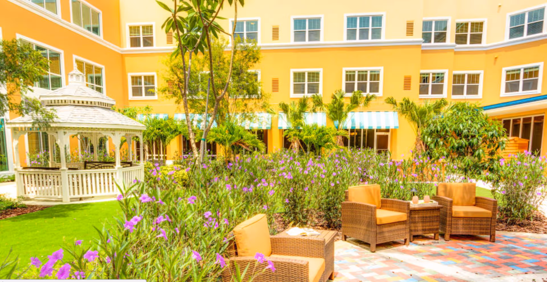 Outdoor courtyard area at Symphony at Delray Beach featuring a white gazebo, lush greenery with purple flowers, and a seating area with wicker chairs and a small table on a colorful tiled patio, surrounded by a yellow building with multiple windows.