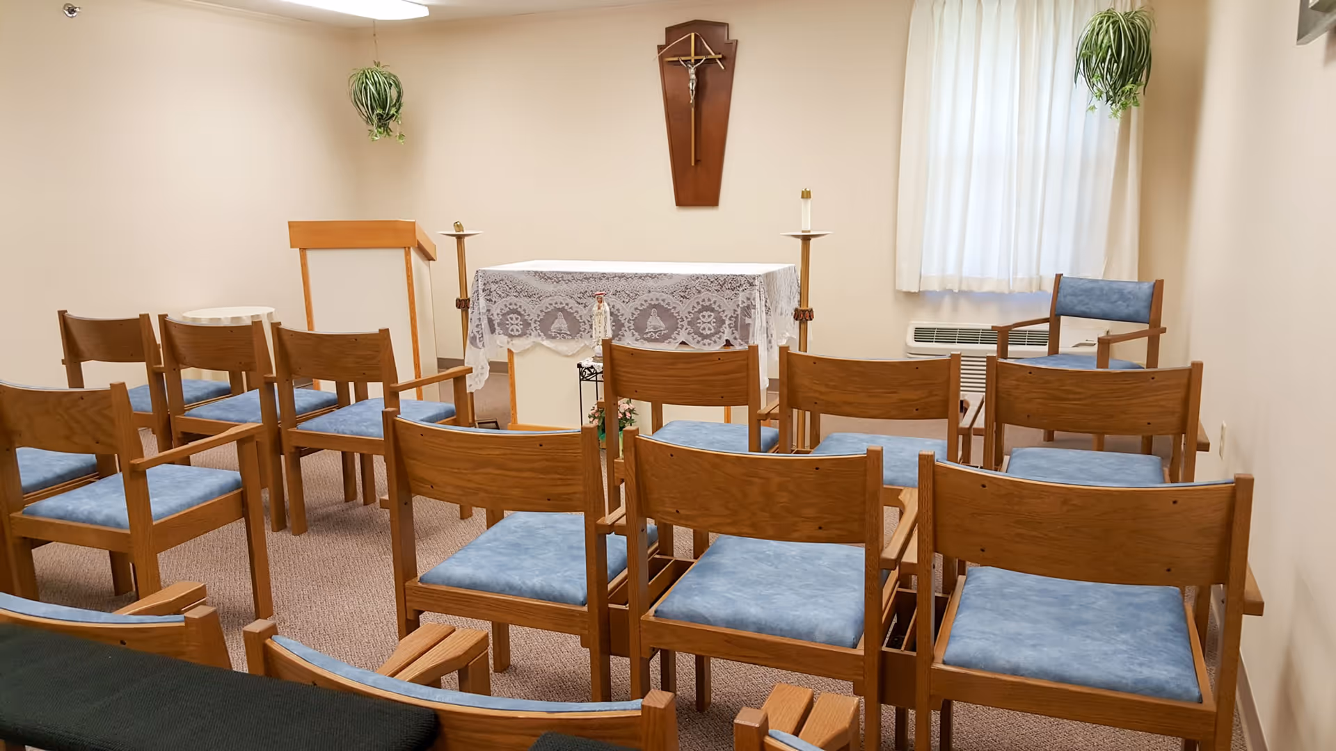 Small chapel room with wooden chairs arranged in rows facing an altar covered with a white lace cloth. A crucifix is mounted on the wall above the altar, and two candles are placed on stands on either side. There are two hanging plants in the corners and a window with white curtains on the right side.