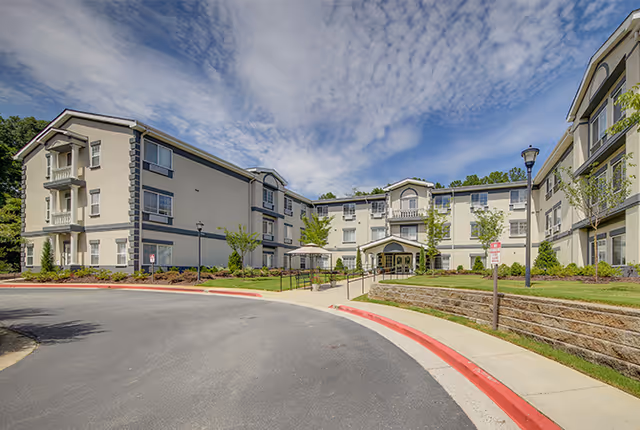Front exterior of a three-story cream-colored senior living building with a curved driveway, landscaped grounds, and main entrance under a partly cloudy blue sky.