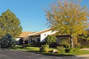Exterior view of a single-story building with a beige roof and light-colored walls, surrounded by green trees and shrubs under a clear blue sky.