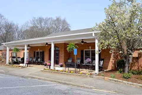 Brick assisted living facility with a covered front porch, rocking chairs, hanging plants, and parking.