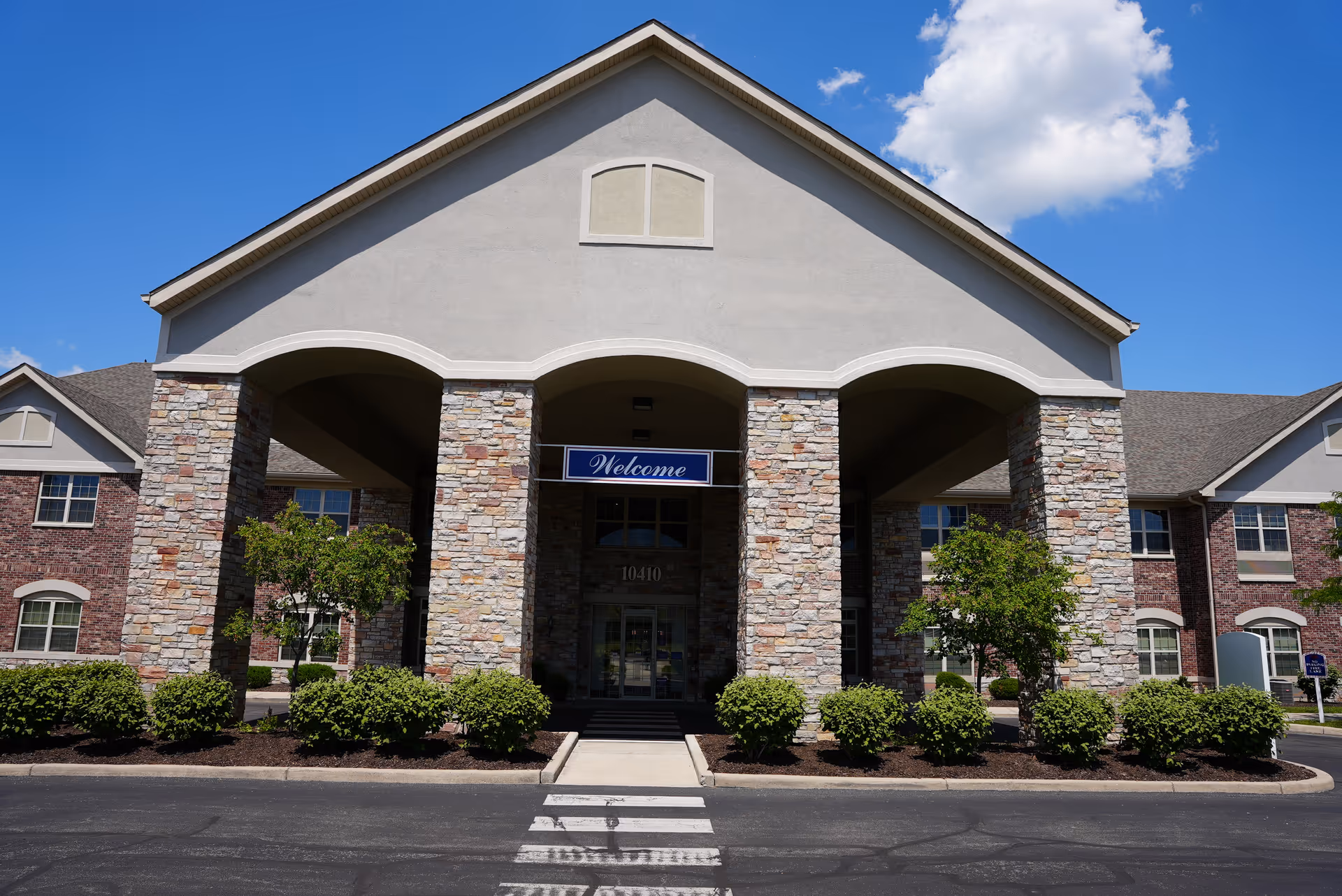 Front entrance of a senior living building with stone columns, a 'Welcome' sign above the doorway and shrubs in front under a blue sky.