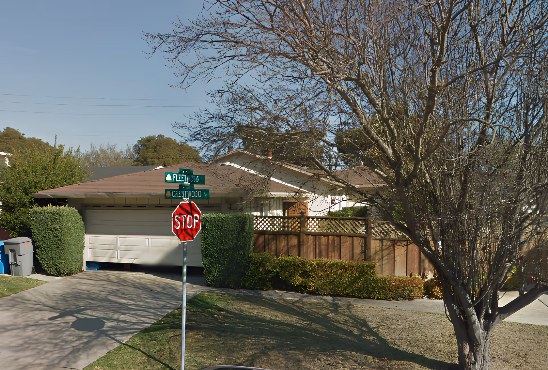 Exterior view of a single-story house with a brown roof and a white garage door. There is a stop sign and street signs at the corner of the driveway. The yard has trimmed bushes, a tree without leaves, and a wooden fence. The sky is clear and blue.