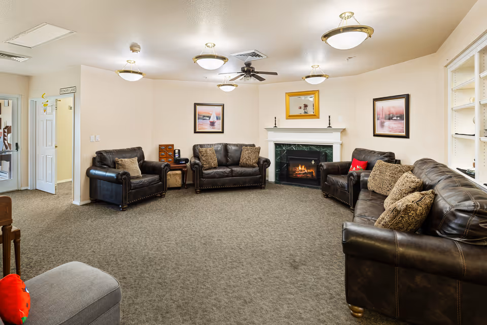 A cozy living room with several dark leather sofas and armchairs arranged around a carpeted floor. There is a fireplace with a fire burning, a ceiling fan, and framed artwork on the walls. The room is softly lit by ceiling lights and has a neutral color scheme.