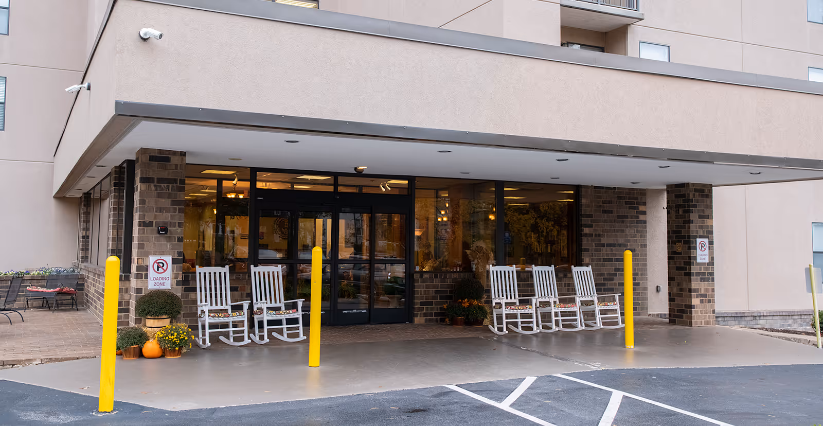 Entrance area of a senior living facility with a covered drop-off zone. There are six white rocking chairs lined up in front of large glass doors, with potted plants and pumpkins placed nearby. The building exterior features beige walls and brick accents. Yellow bollards are positioned to protect the walkway, and signs indicate a loading zone with no parking allowed.