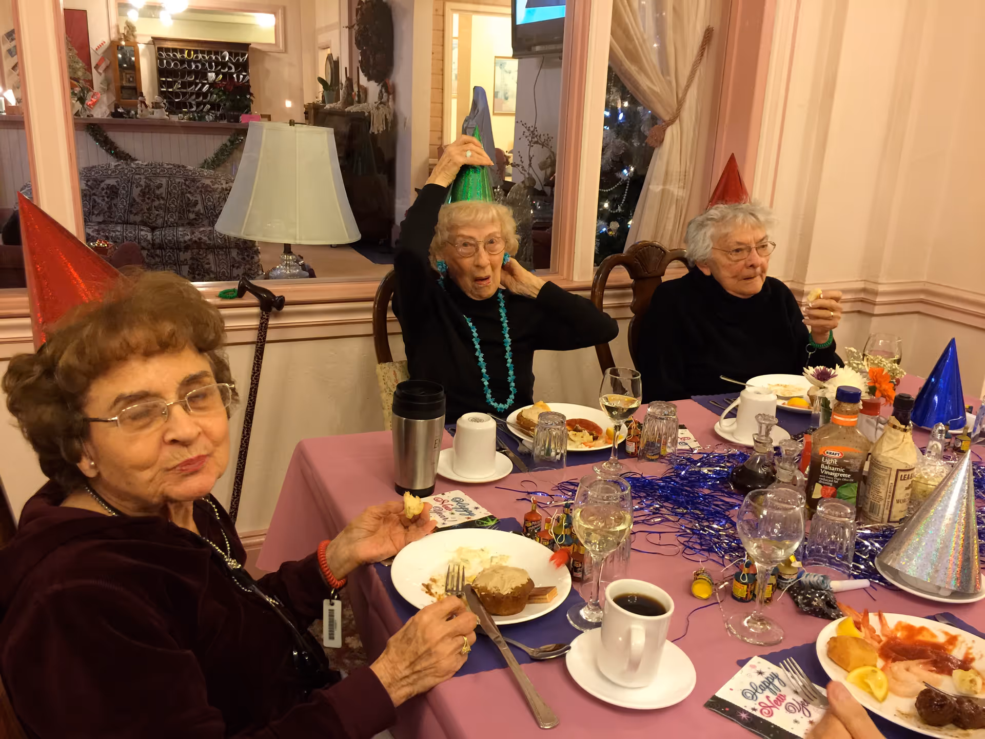 Three elderly women wearing colorful party hats sitting around a table with food and drinks, celebrating an event in a decorated room with a lamp and a mirror in the background.