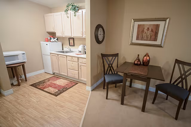 A small kitchen area with light wood cabinets, a white refrigerator, a microwave on a small table, and a sink. Adjacent to the kitchen is a dining area with a dark wooden table, two chairs with dark cushions, two decorative vases on the table, and a framed floral picture on the wall.