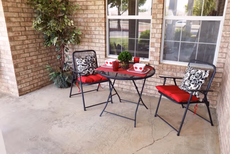 A small outdoor patio area with two black metal chairs featuring red seat cushions and black-and-white patterned back cushions. Between the chairs is a round black metal table with a red placemat, two white bowls with red dots, two red cups, and a small green potted plant. The patio is surrounded by brick walls and has two windows in the background. There is also a tall green potted plant in the corner.