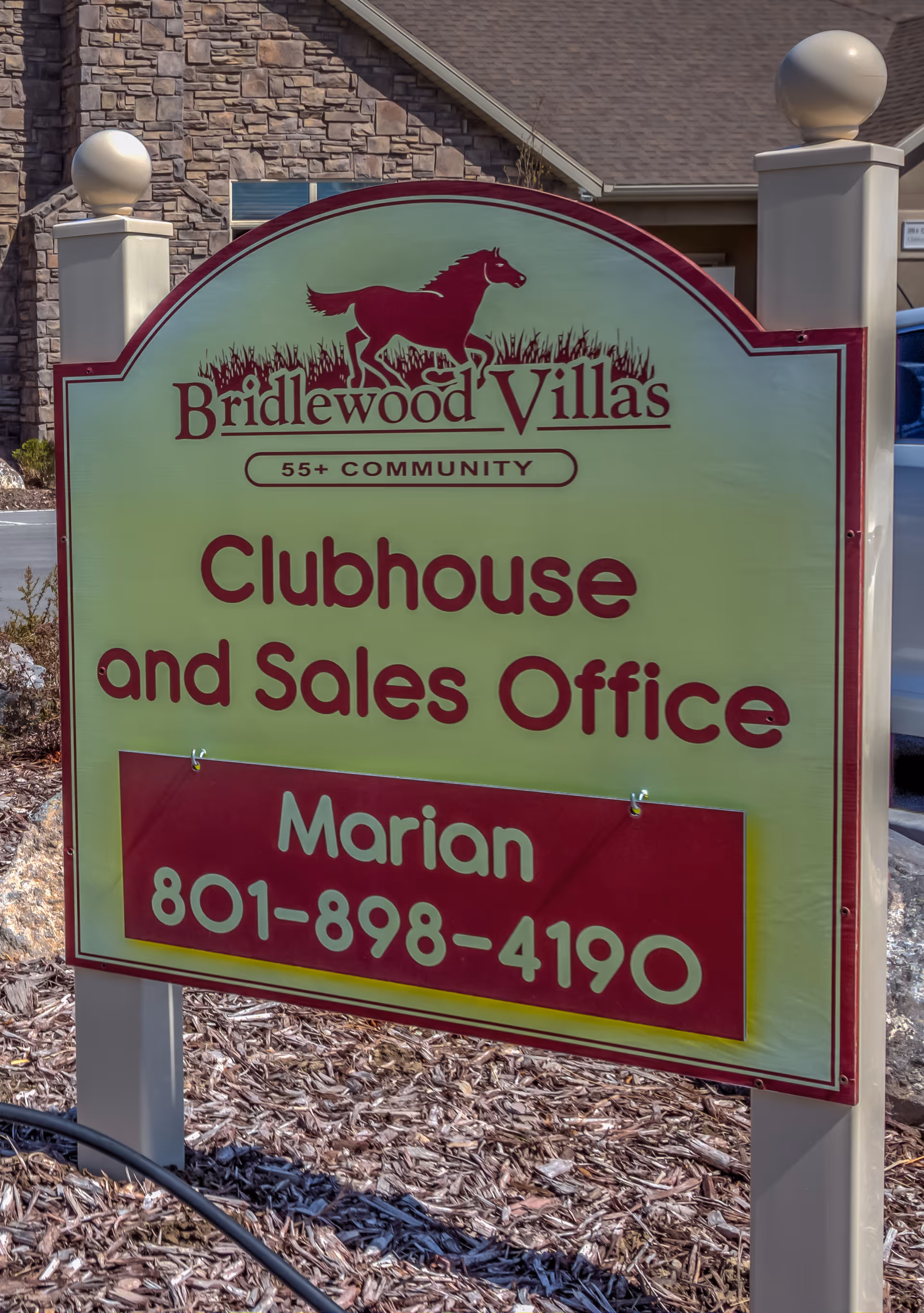 Outdoor sign for Bridlewood Villas 55+ community indicating the Clubhouse and Sales Office with contact name Marian and phone number 801-898-4190. The sign has a horse logo and is mounted on two posts with a building and landscaping visible in the background.