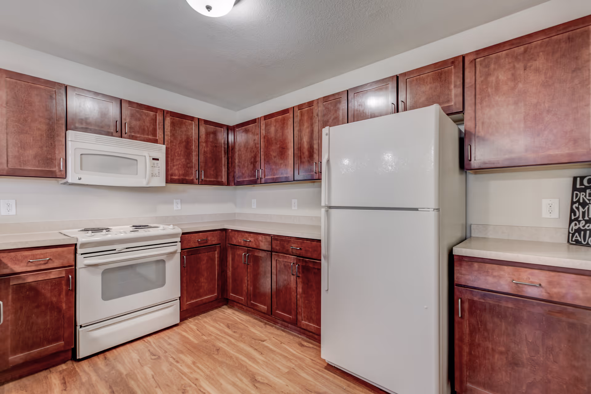 A kitchen with wooden cabinets, a white refrigerator, a white stove with oven, and a white microwave mounted above the stove. The countertops are light-colored, and the floor has a wood finish. There is a small decorative sign on the right countertop.