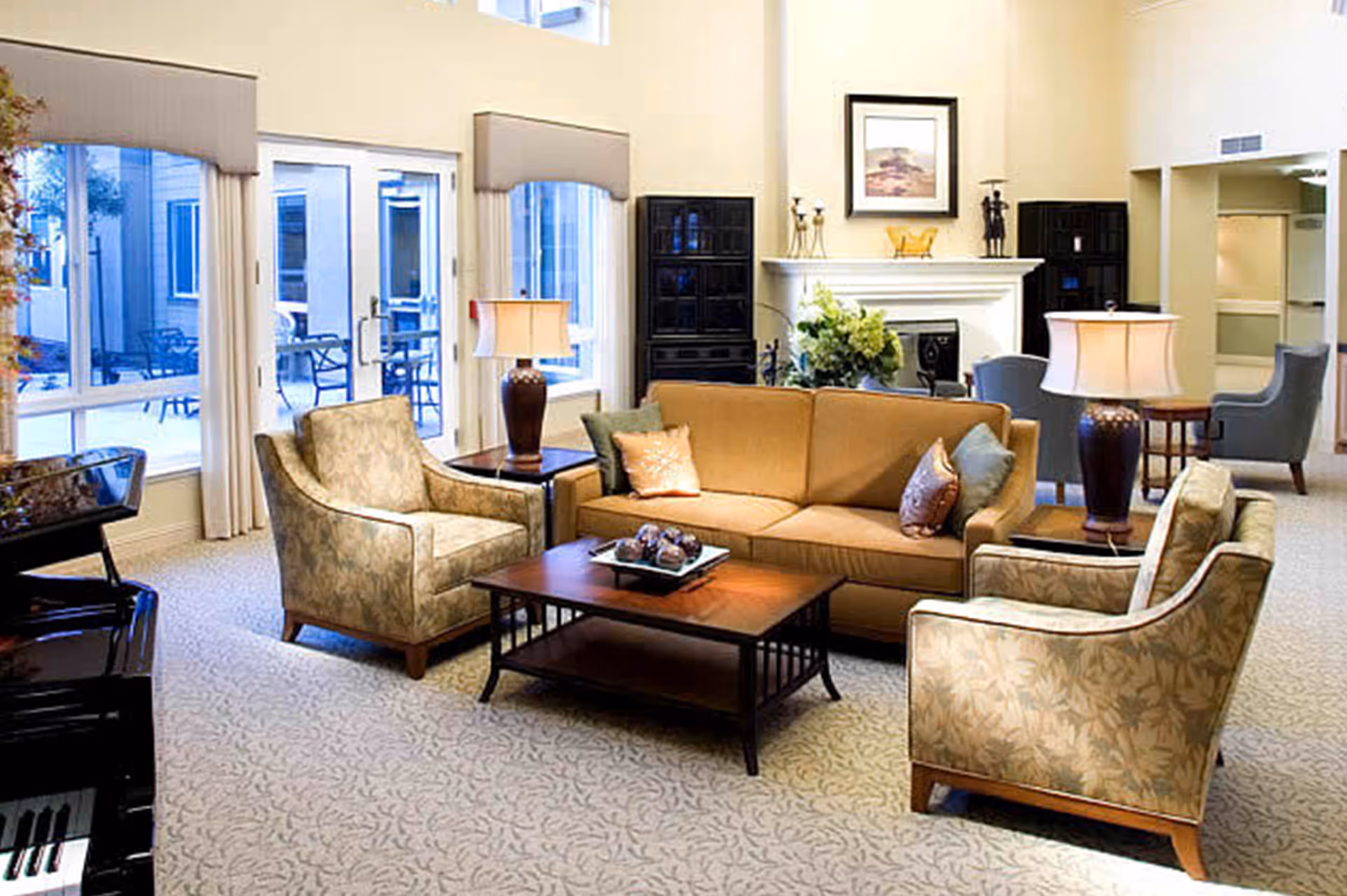 Bright communal living room with a sofa, upholstered armchairs, coffee table, lamps, and a fireplace in front of large windows.