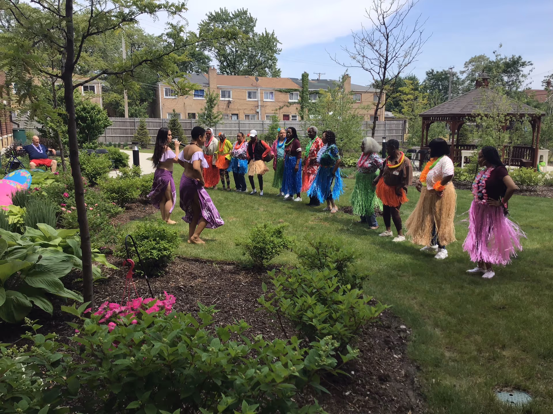 People in colorful hula skirts dancing on the lawn of a garden courtyard with plants and a gazebo in the background.