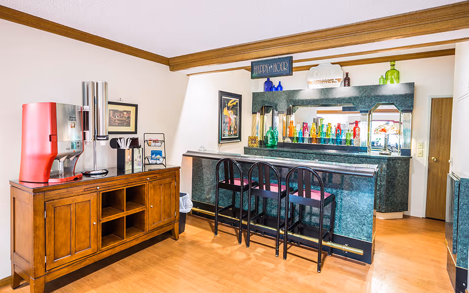 A small bar area with a green marble countertop and three black bar stools with red cushions. Behind the bar is a large mirror with colorful glass bottles displayed on the counter. To the left, there is a wooden cabinet with a red beverage dispenser and some utensils. The room has light-colored walls, wooden trim, and a wooden floor. There are framed pictures on the walls and a sign above the bar that reads 'HAPPY HOUR'.