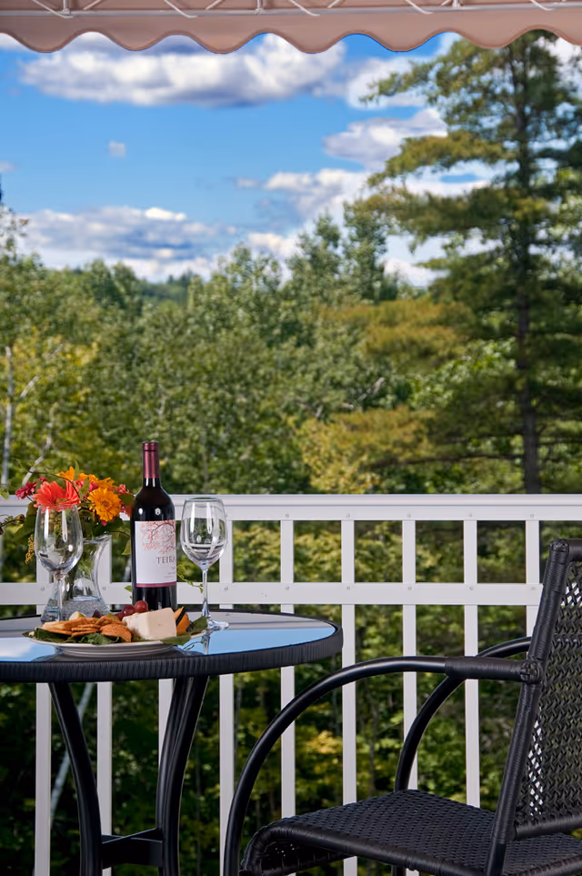 A balcony table set with a bottle of wine, two wine glasses, a plate of snacks and a vase of flowers overlooking a tree-filled landscape.