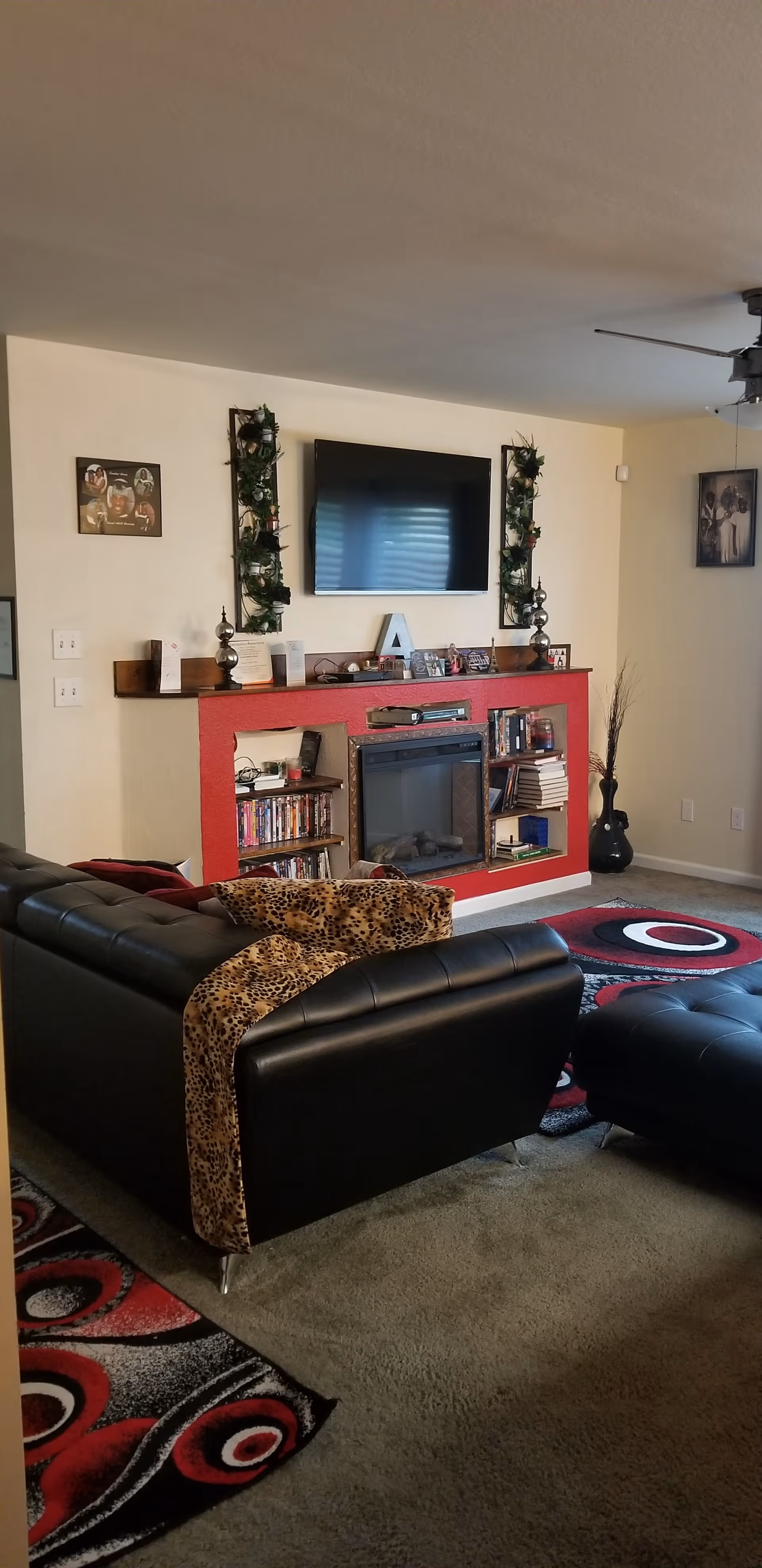 Living room with a black leather sectional and ottoman facing a wall-mounted TV above a red-accent fireplace with shelves and decorative items.