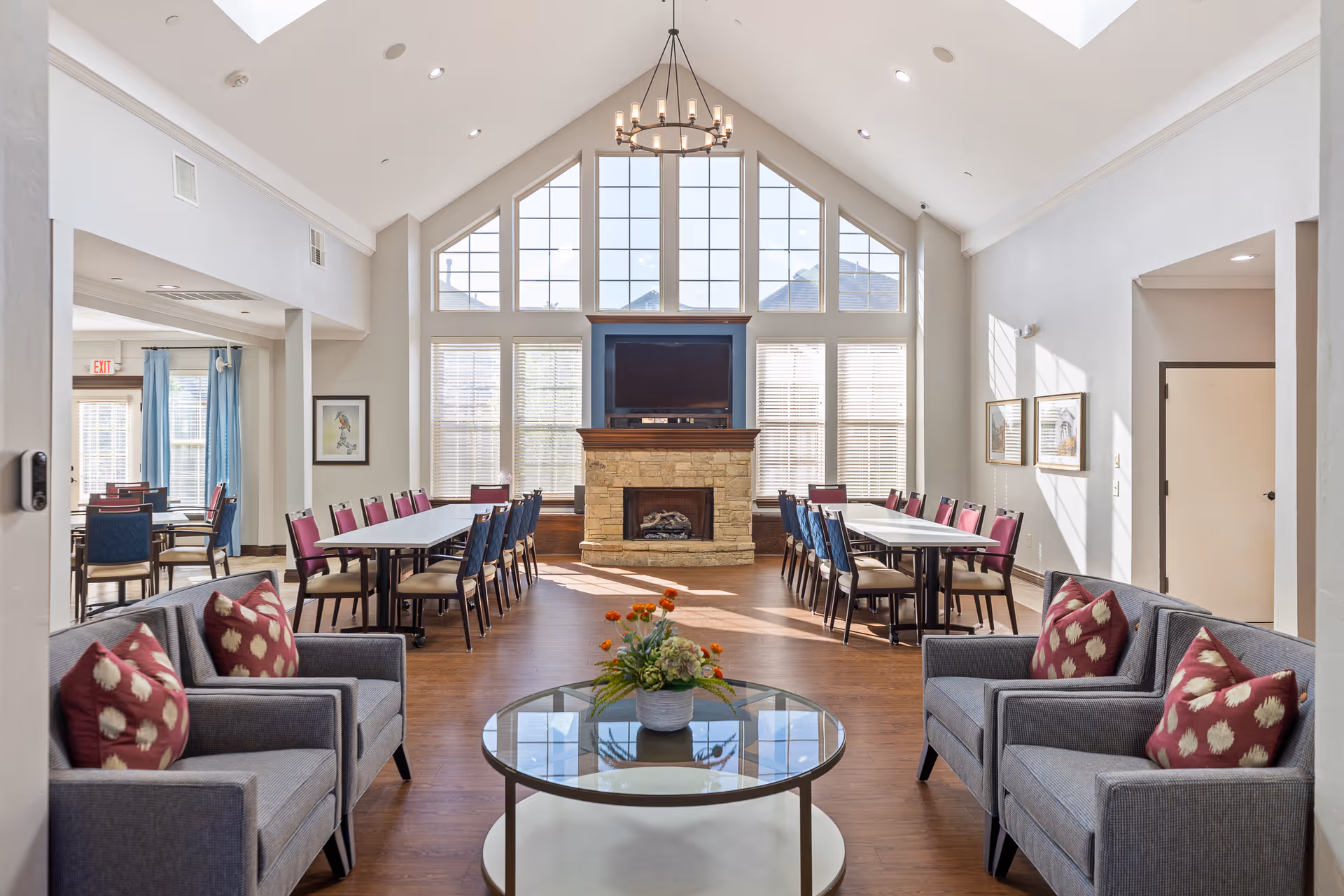 A bright and spacious common area with high vaulted ceilings and large windows letting in natural light. The room features two long dining tables with chairs, a stone fireplace with a TV mounted above it, and two gray sofas with red patterned pillows facing a round glass coffee table with a flower arrangement. The walls are painted white, and there are framed pictures on the right wall.