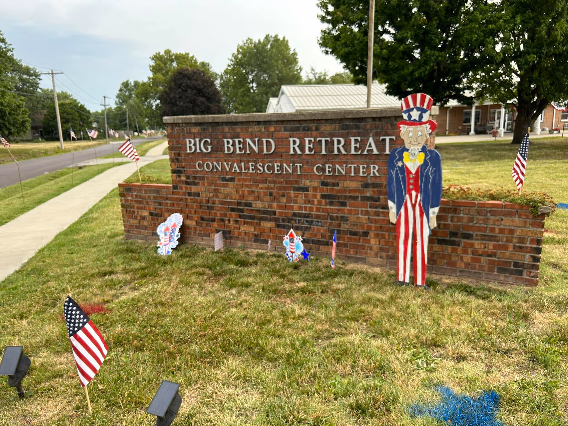 A brick sign for Big Bend Retreat Convalescent Center with small American flags and patriotic decorations, including a wooden cutout of Uncle Sam, on a grassy area beside a sidewalk and road.