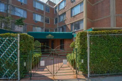Brick building entrance with gated walkway and green hedges leading to a covered doorway.