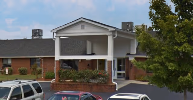 Front exterior view of a single-story brick building with a covered entrance and several parked cars in front. There are some bushes and a tree near the entrance under a partly cloudy sky.