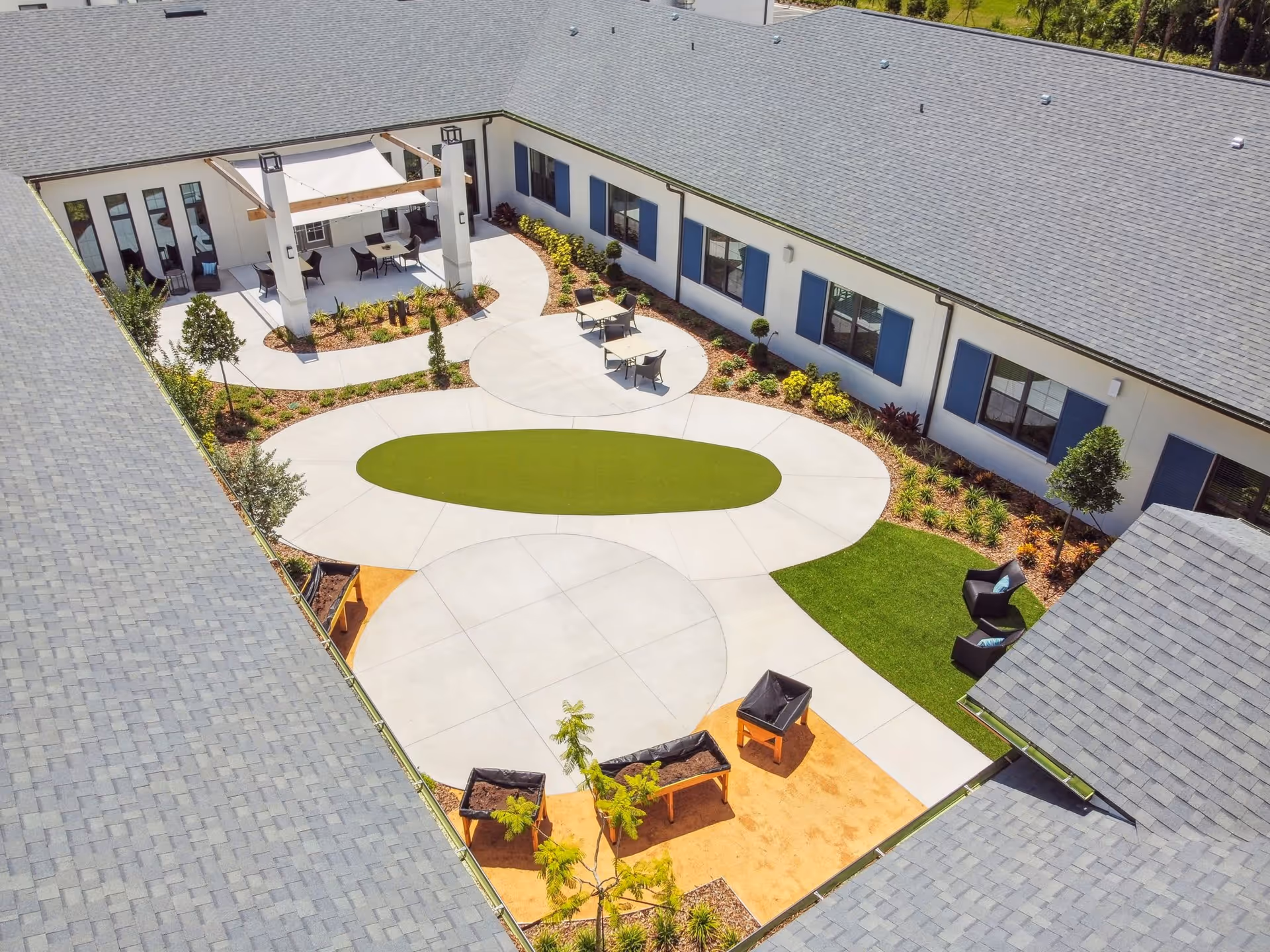 Aerial view of a senior living facility courtyard with a paved walking path, green lawn areas, several tables and chairs, a shaded seating area, and raised garden beds surrounded by a building with blue window shutters.