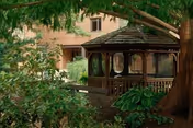 A wooden gazebo surrounded by lush green plants and trees in an outdoor garden area with a building visible in the background.