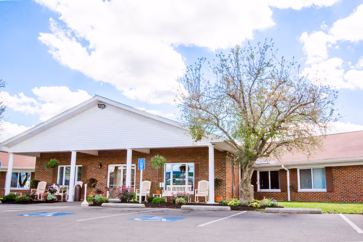 Front exterior view of a single-story brick building with a white gabled roof and a covered entrance supported by white columns. There are several white rocking chairs and potted plants near the entrance. A large tree with sparse leaves stands to the right of the entrance. The parking lot in front has marked handicap parking spaces. The sky is partly cloudy with blue patches.