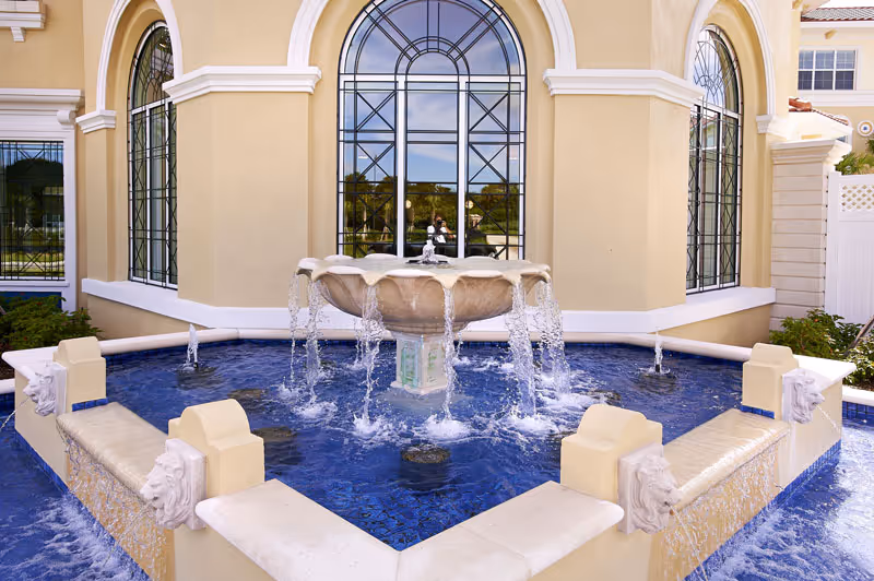 Outdoor water fountain with multiple streams of water flowing into a blue-tiled basin, situated in front of a beige building with large arched windows featuring decorative black metal grilles.