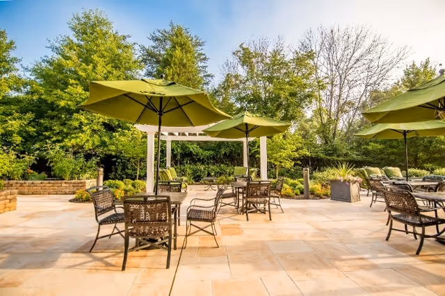 Sunlit outdoor patio with tables, chairs, and green umbrellas surrounded by trees and landscaping.