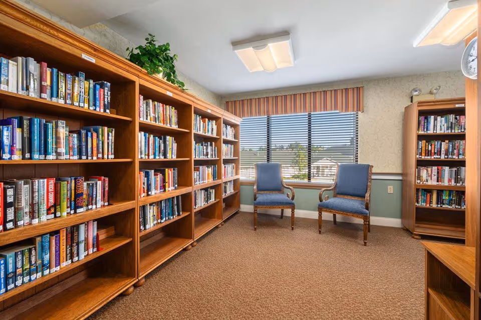 Cozy library room with wooden bookshelves filled with books, two blue upholstered chairs, and a window with blinds.