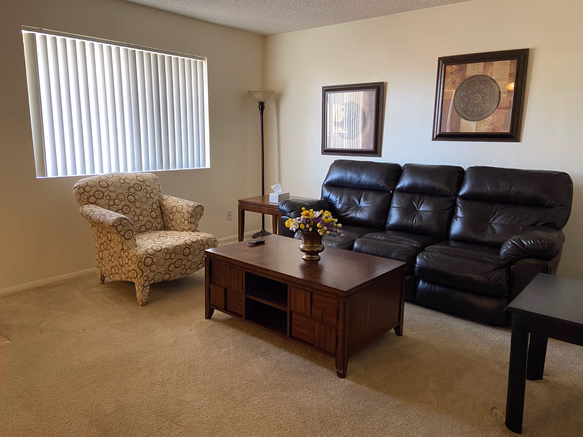 A senior living facility living room with a patterned armchair, a dark brown leather sofa, a wooden coffee table with a flower vase, a side table with a tissue box, a floor lamp, and two framed decorative wall art pieces. A window with vertical blinds lets in natural light.
