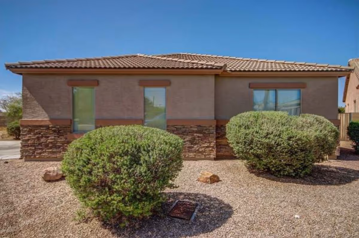 Exterior view of a single-story building with a tiled roof, two windows with blinds, and a stone and stucco facade. In front of the building are two large green bushes and gravel landscaping under a clear blue sky.