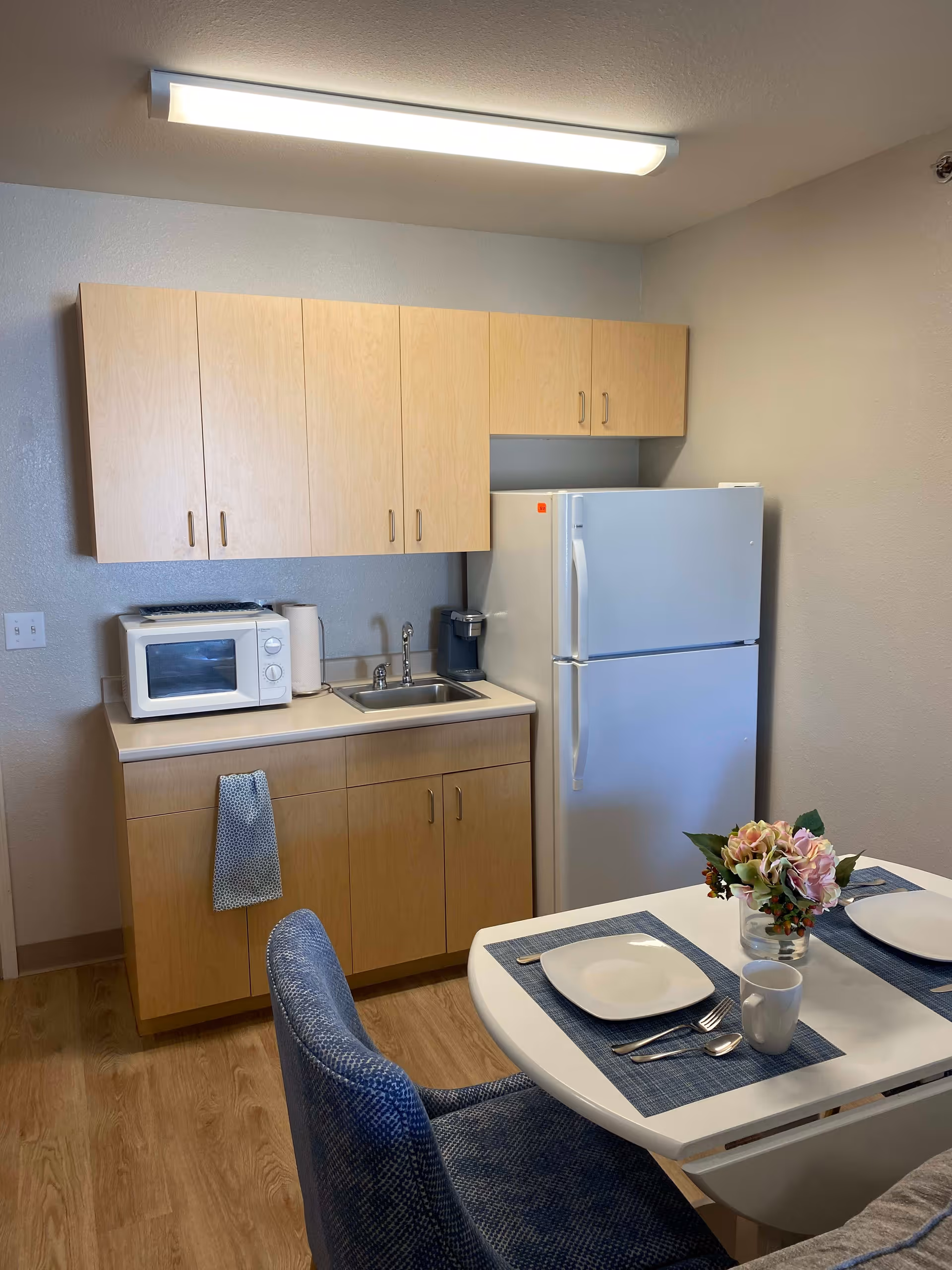 A small kitchen area with light wood cabinets, a white microwave, a paper towel roll, a sink, a coffee maker, and a white refrigerator. In the foreground, there is a dining table set with two place settings including plates, utensils, a mug, and a vase with flowers. Two blue upholstered chairs are positioned at the table.