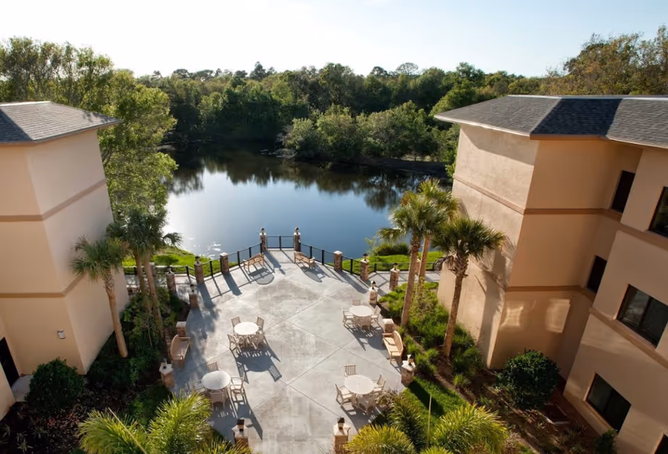 Outdoor patio area between two beige buildings with round tables and chairs, palm trees, and a view of a calm pond surrounded by trees in the background.