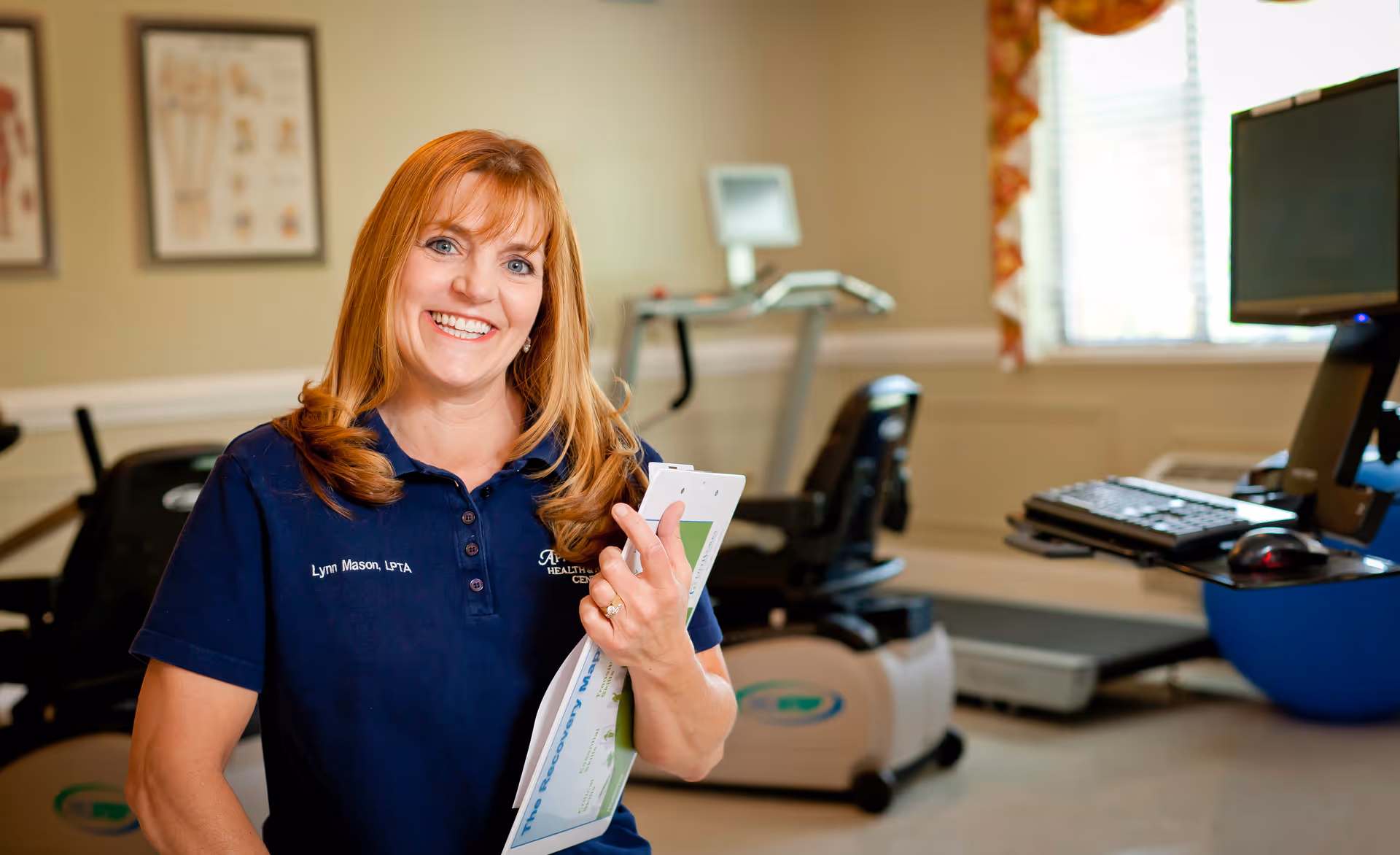 A smiling woman with red hair wearing a navy blue polo shirt with the name Lynn Mason, LPTA, holding papers in a room with exercise equipment and a computer workstation.