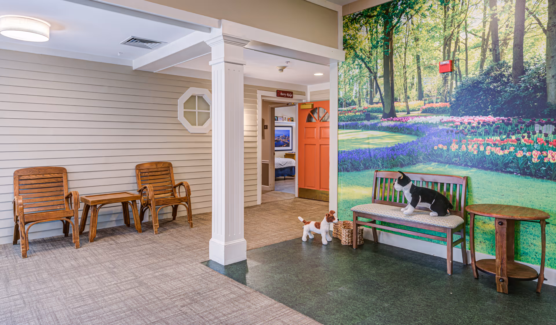 Indoor seating area with two wooden chairs and a small table on the left, a white column in the center, and a bench with a cushion on the right. Two stuffed animal dogs are placed near the bench. The wall behind the bench features a large mural of a garden with trees and flowers. An open doorway with an orange door leads to another room with a bed visible inside.