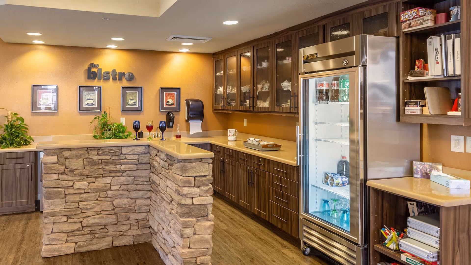Interior view of a bistro area with a stone-faced counter, wooden cabinets with glass doors, a glass-front refrigerator stocked with beverages, and shelves holding various items. The wall behind the counter has framed pictures and the words 'the bistro'. There are also some plants and a few glasses on the counter.