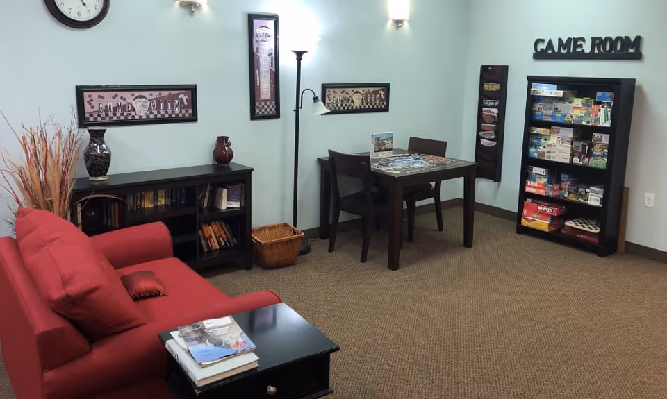 A cozy game room with a red couch, a black side table with books, a bookshelf filled with books, a small table with two chairs, a floor lamp, and a cabinet stocked with various board games. The walls are light blue with framed game-themed artwork and a clock. The words 'GAME ROOM' are displayed on the wall above the game cabinet.