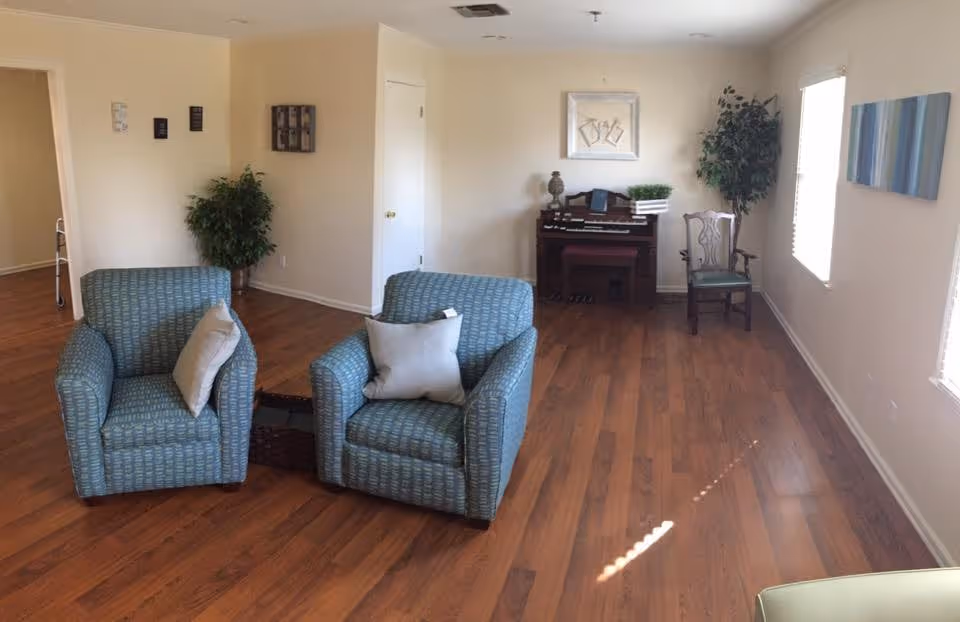A bright living room with wooden flooring featuring two blue patterned armchairs with light-colored pillows, a small wooden table between them, a wooden chair, a potted plant, a framed picture on the wall, and a small organ or piano against the far wall.