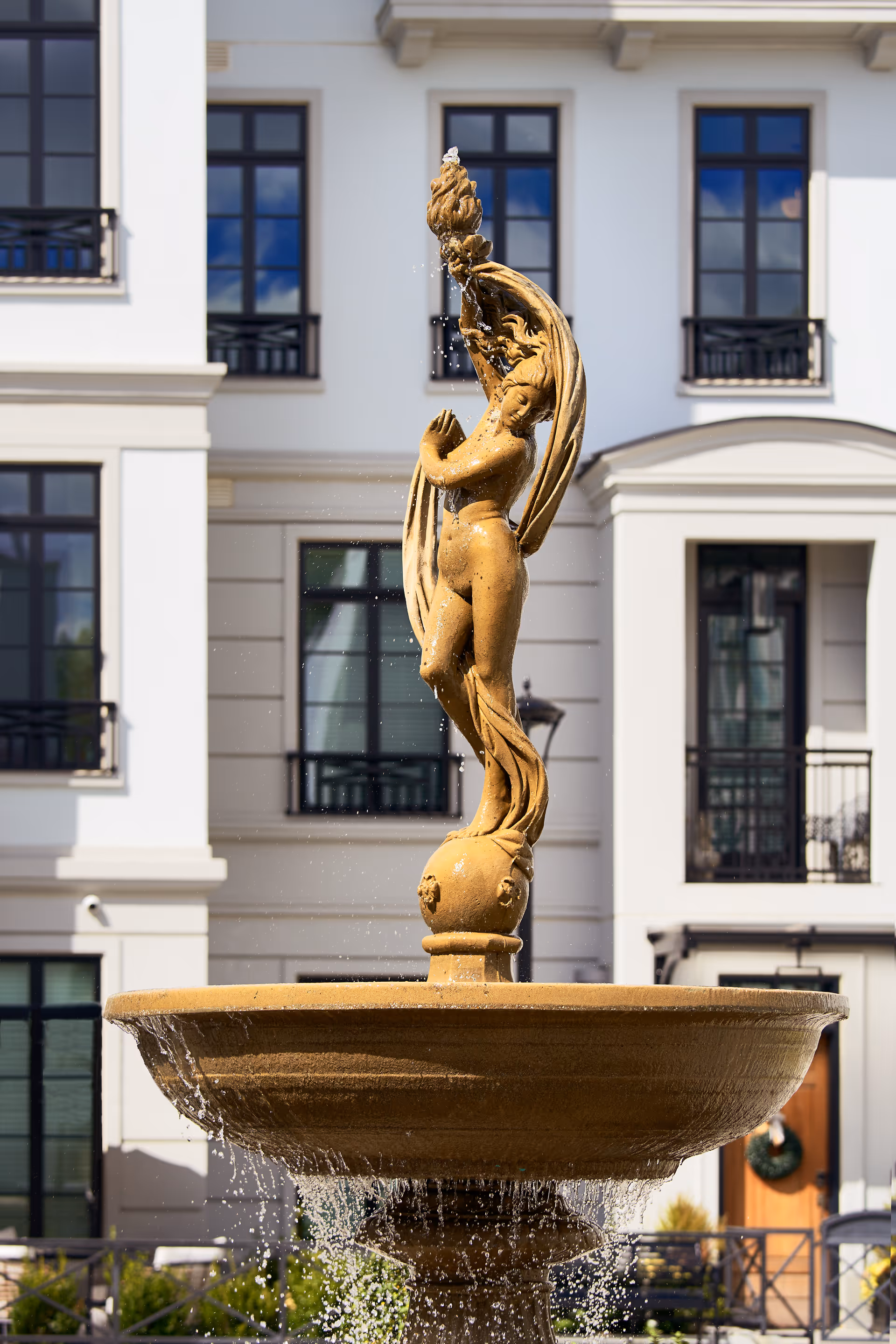 Ornate stone fountain topped by a sculpted female figure with water cascading in front of a white multi-story building facade.