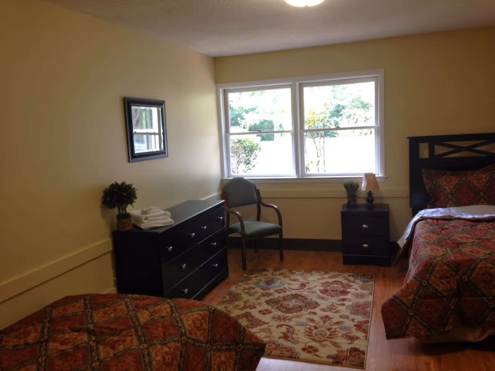 A bedroom with two beds covered in red and orange patterned bedspreads. There is a black dresser with a small potted plant and folded towels on top, a green upholstered chair near a window, and a black nightstand with a small lamp and decorative plant. A colorful area rug is on the wooden floor.