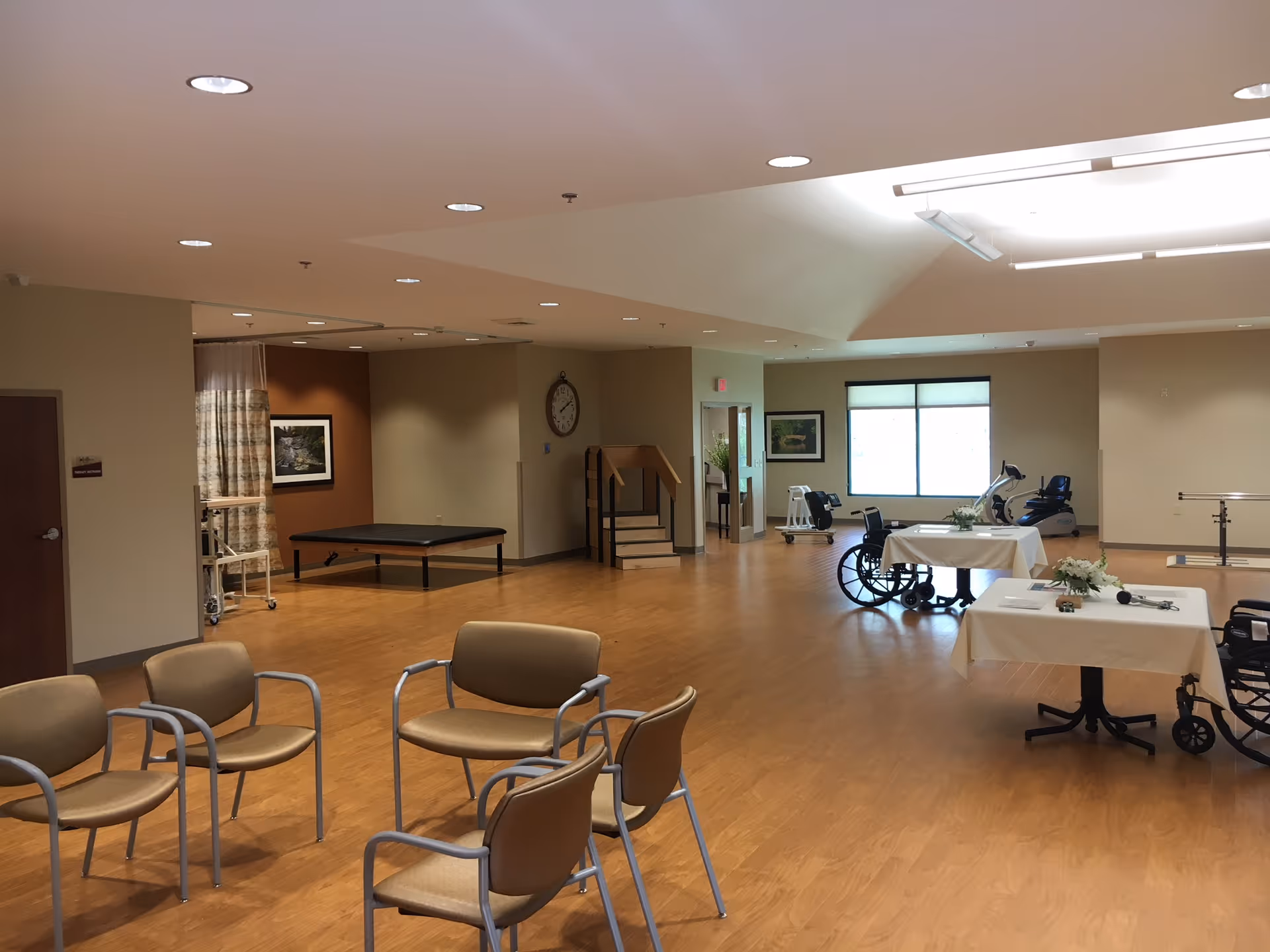 A spacious room with wooden flooring and beige walls, featuring several chairs arranged in a circle, tables covered with white cloths, wheelchairs, exercise equipment, and a small set of stairs. The room is well-lit with ceiling lights and natural light from a window.