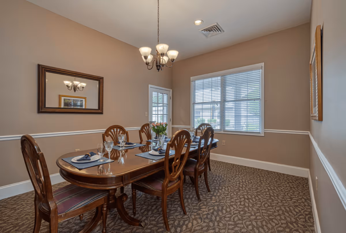A dining room with a wooden table set for six people, featuring glassware, napkins, and a vase of flowers. The room has beige walls with white trim, a patterned carpet, a window with blinds, a chandelier hanging from the ceiling, and a large mirror on one wall.