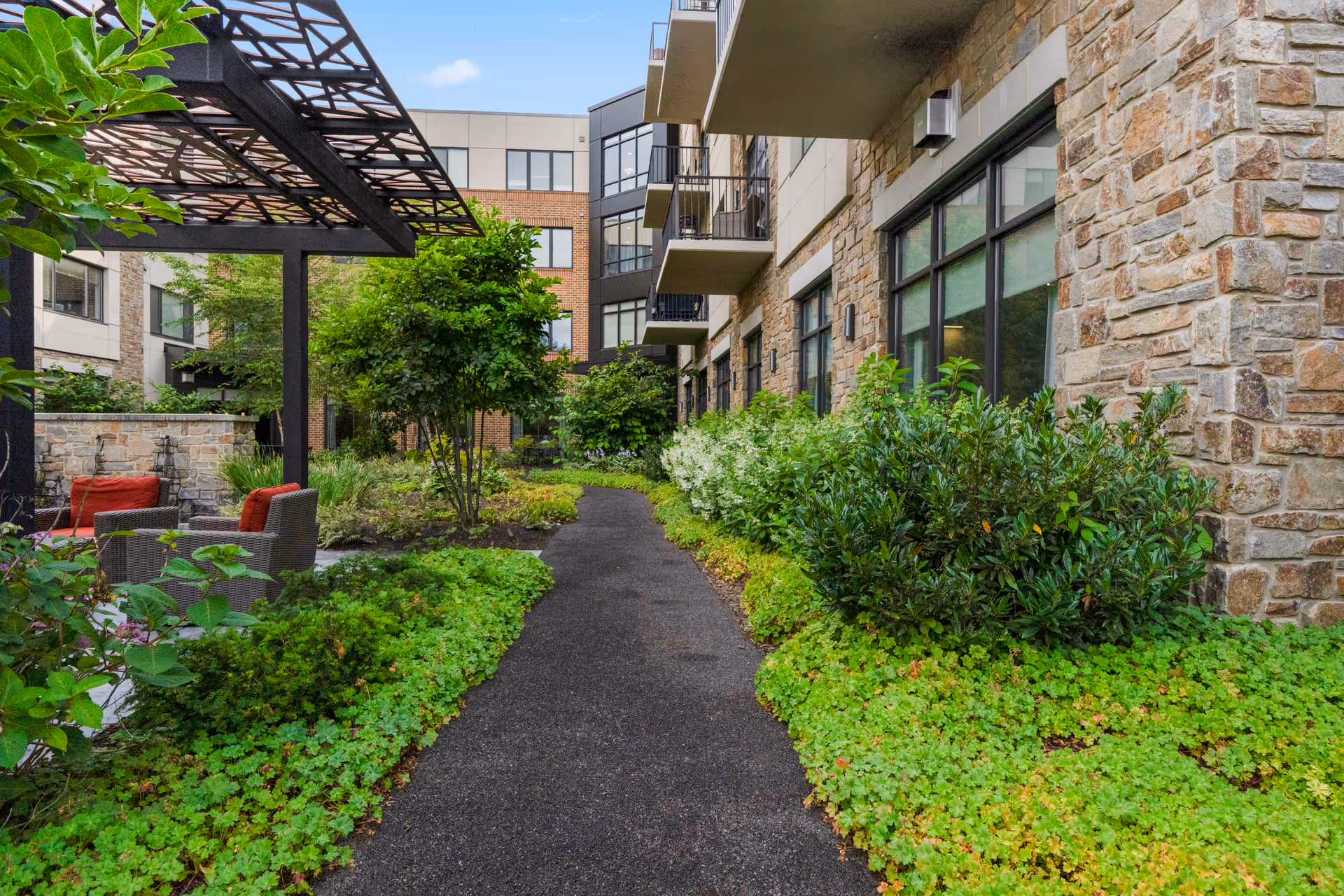 Outdoor garden area at Eagleview Landing featuring a paved walkway surrounded by lush green plants and bushes, with a stone building on the right and a pergola with seating on the left.