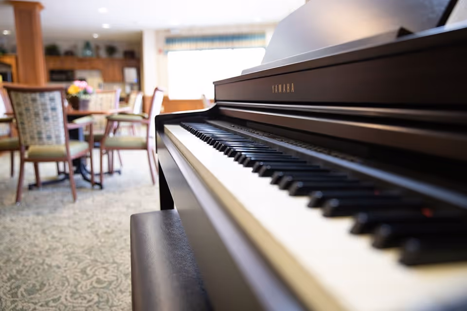 Close-up view of a Yamaha piano keyboard in a well-lit room with carpeted floor. In the background, there are tables and chairs arranged in a common area with a window letting in natural light.