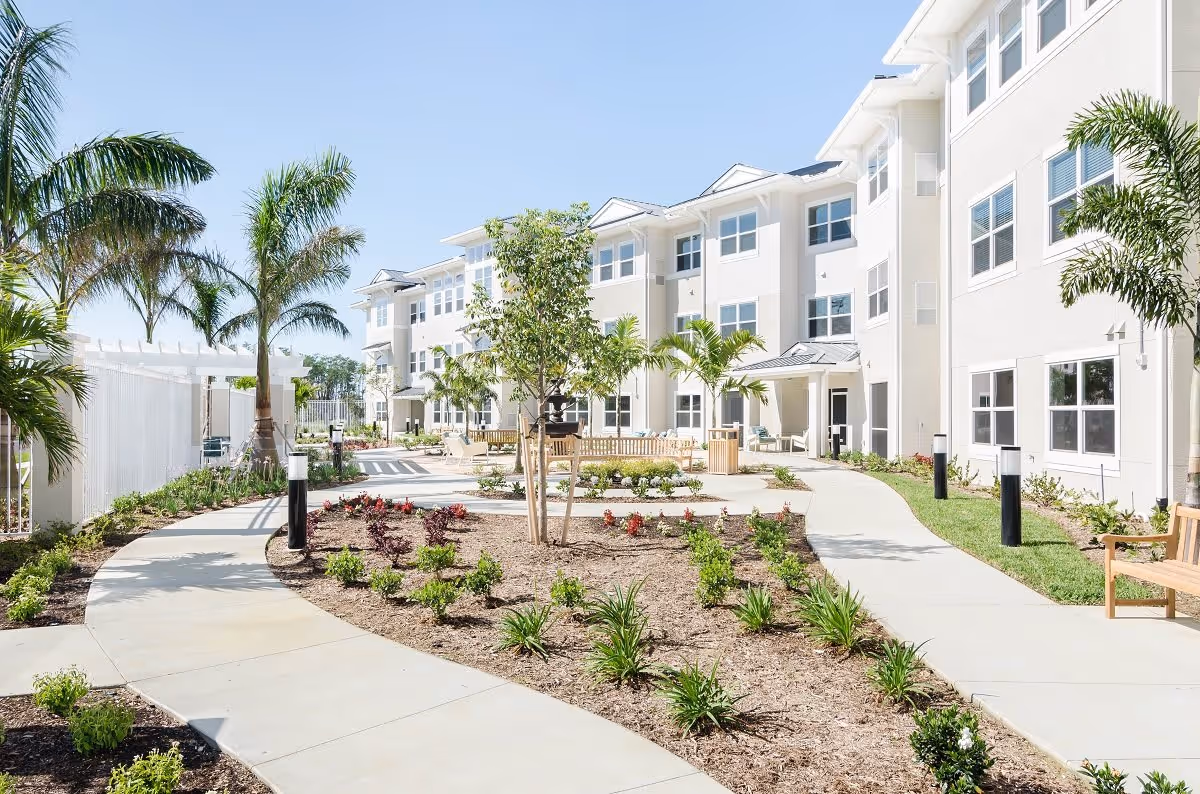 Outdoor courtyard area of a senior living facility with paved walkways, landscaped garden beds, palm trees, benches, and a three-story building in the background under a clear blue sky.